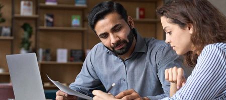 Two multiethnic professionals colleagues working together with laptop and papers in office. Indian male mentor and latin female young professional sitting in creative office space.