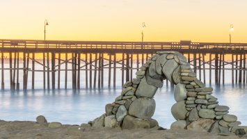 Beach rocks delicately stacked into an arch with the lights of Ventura pier glowing in the lamps after dawn.